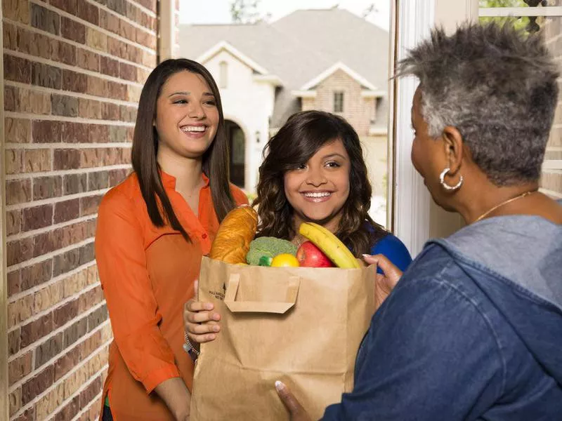 Volunteers: Young adults bring groceries to senior woman.