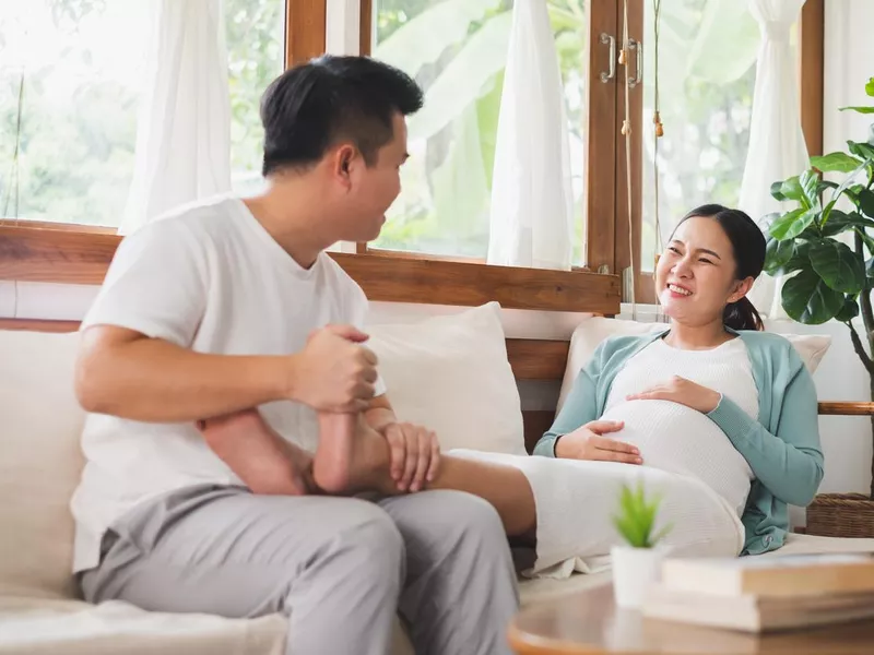 Asian young husband massaging pregnant wife feet at home, happy family