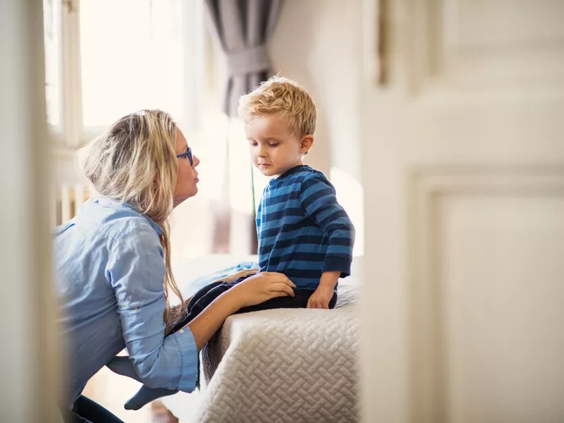 A young mother talking to her toddler son inside in a bedroom.