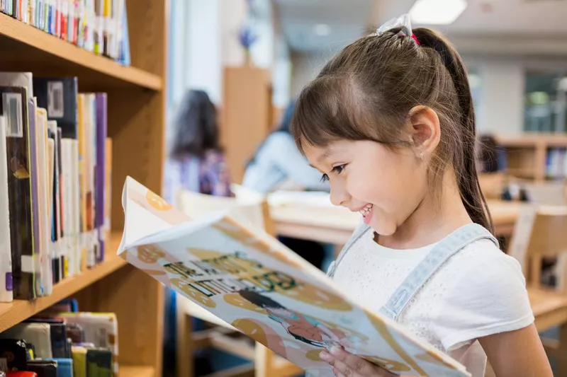 Happy little girl reads book in school library