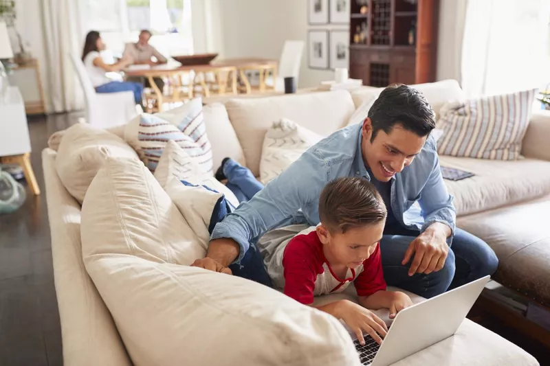 First grade boy lying on sofa using laptop with his dad. Mum and grandad at the table in the background