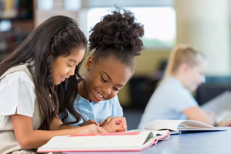 Smiling and cheerful schoolgirls reading a book together at school