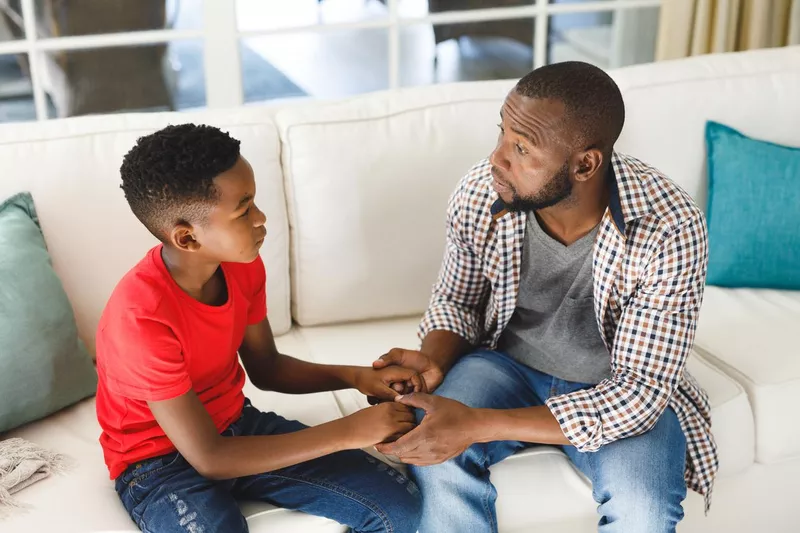 Father and son sitting on couch in living room talking and holding hands