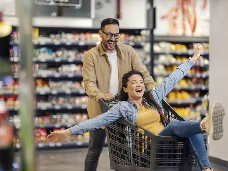 A happy man drives his woman in shopping cart in supermarket.